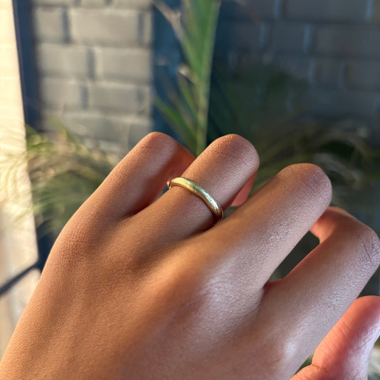 A close-up of a hand wearing a simple textured gold band ring, set against a blurred background of a gray brick wall and green foliage.