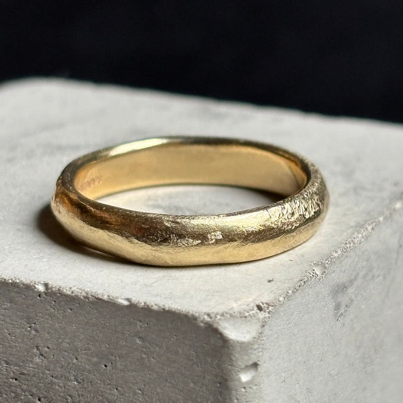 A textured yellow gold band ring resting on a light gray concrete block against a dark background.