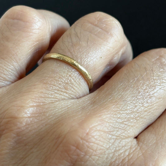 A close-up of a hand wearing a simple textured yellow gold band ring against a dark background.