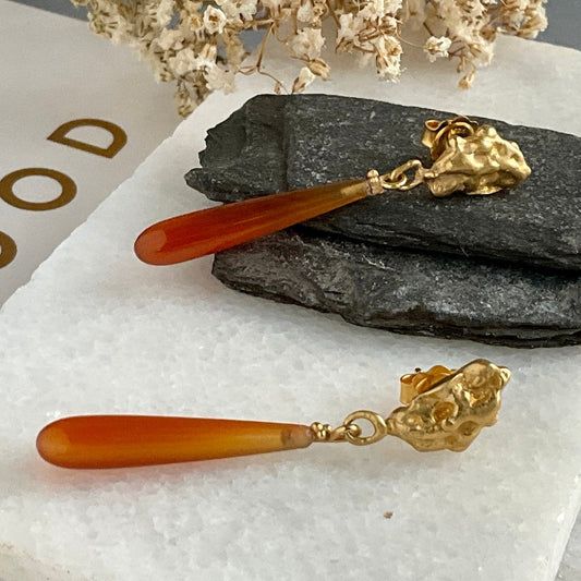 Pair of earrings with gold accents and orange stones on a textured surface with rocks and dried flowers.