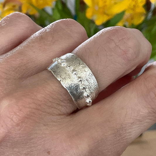 Silver ring with textured design worn on a finger, with yellow flowers in the background