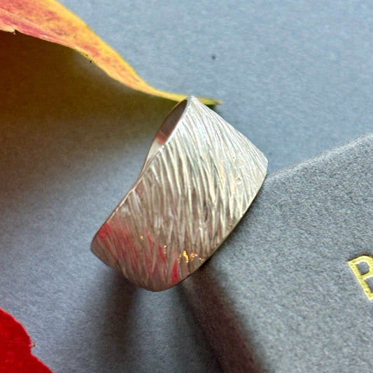 A silver men's ring with a textured surface, displayed on a grey card, with red leaf accents in the background.