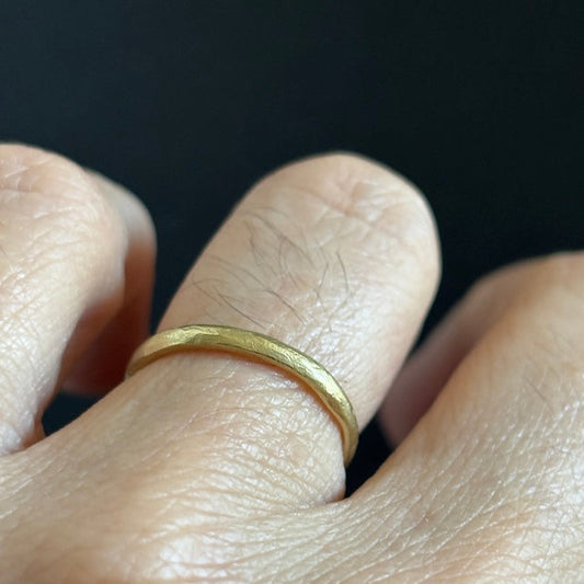 A close-up of a hand wearing a simple, thin yellow gold band ring on one of the fingers, set against a solid black background.