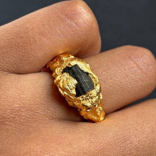 A gold ring with a textured, organic-shaped setting holding a dark green banded stone, shown worn on a person's finger.