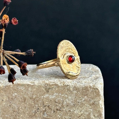 Gold ring with a red gemstone on a stone surface with dried flowers.