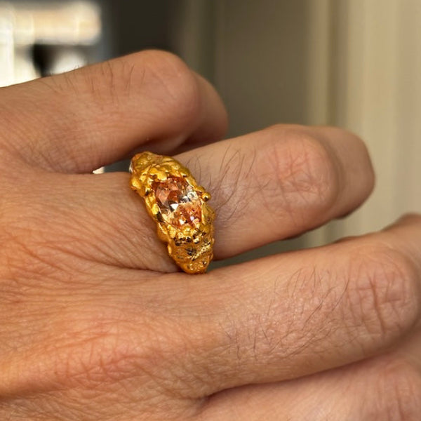A close-up of a hand wearing a textured gold ring with a central peach-colored gemstone.