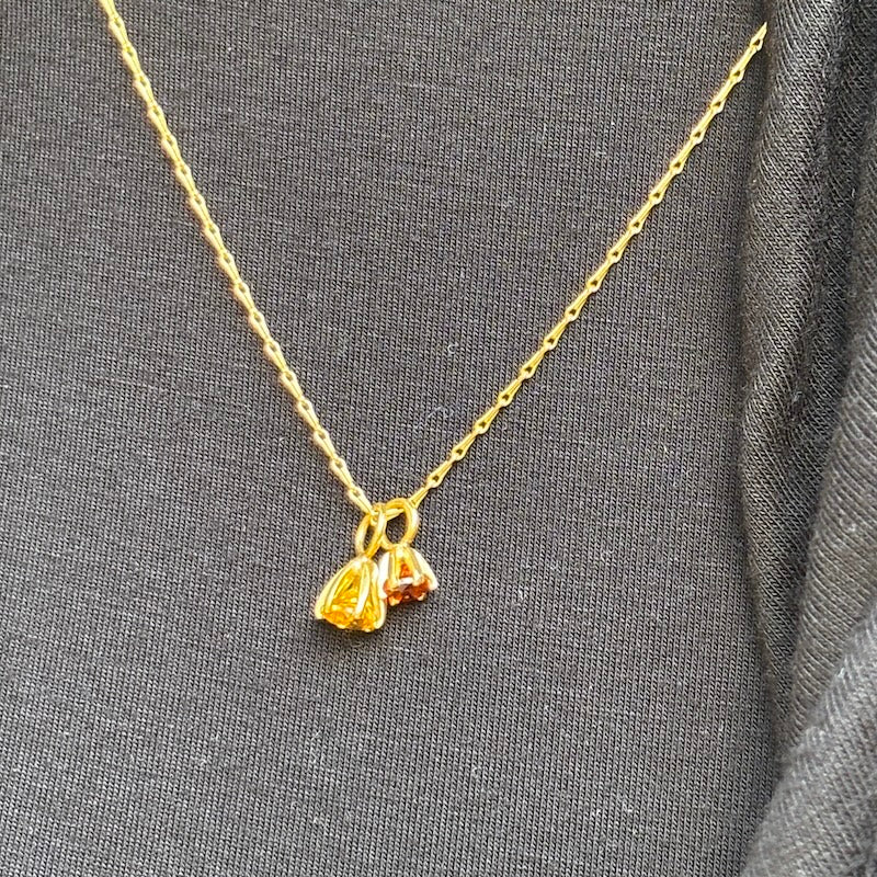 A delicate gold chain necklace with a small pendant featuring two small flower-like charms, one yellow gold and one with a warm orange-red center, displayed on a textured dark gray fabric background.