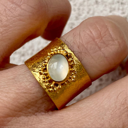 A person's hand displaying a gold-colored open band ring with a white round gemstone set in a gold-colored bezel on top.
