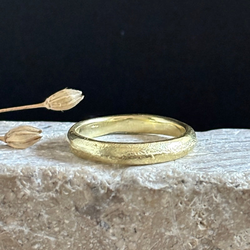 A textured yellow gold band ring resting on a light stone surface with dried flower buds against a dark background.