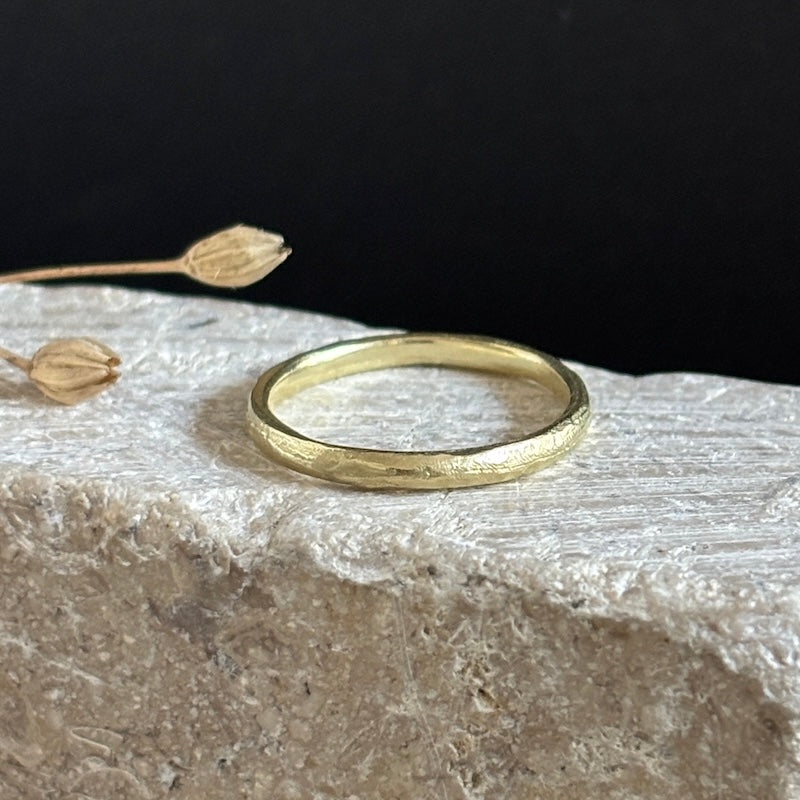 A textured yellow gold band ring resting on a rough white stone surface with dried flower buds against a black background.