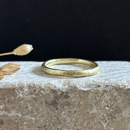 A textured yellow gold band ring resting on a rough stone surface with dried flower buds against a dark background.