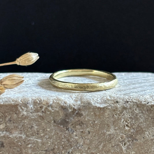 A textured yellow gold band ring resting on a rough stone surface with dried flower buds against a dark background.