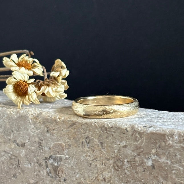 A textured gold band ring resting on a light stone ledge next to dried white daisy flowers, set against a dark black background.