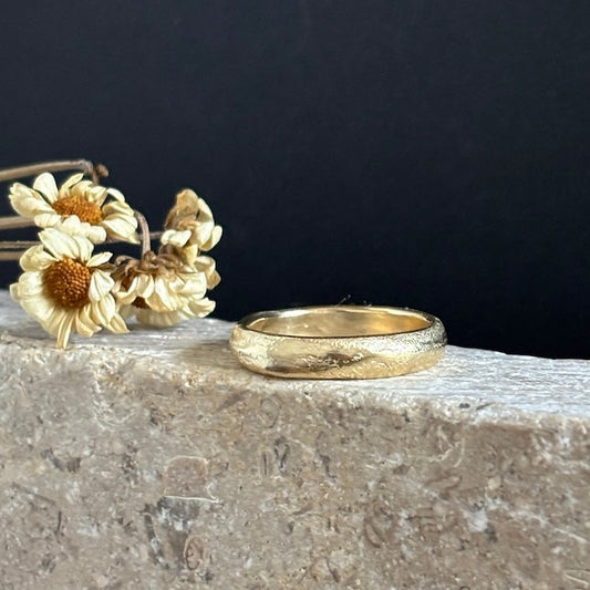 A textured gold band ring resting on a light stone ledge next to dried white daisy flowers, set against a dark black background.