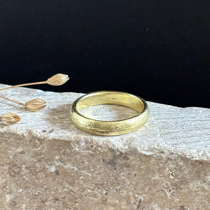 A textured yellow gold band ring resting on a light-colored stone slab with dried flower buds against a solid black background.