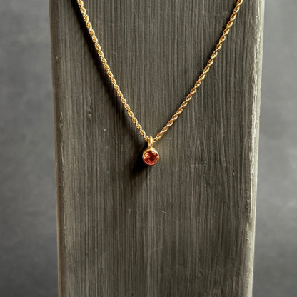 A delicate gold chain necklace with a small round amber-brown gemstone pendant, displayed on a vertical gray wooden stand against a dark gray background.