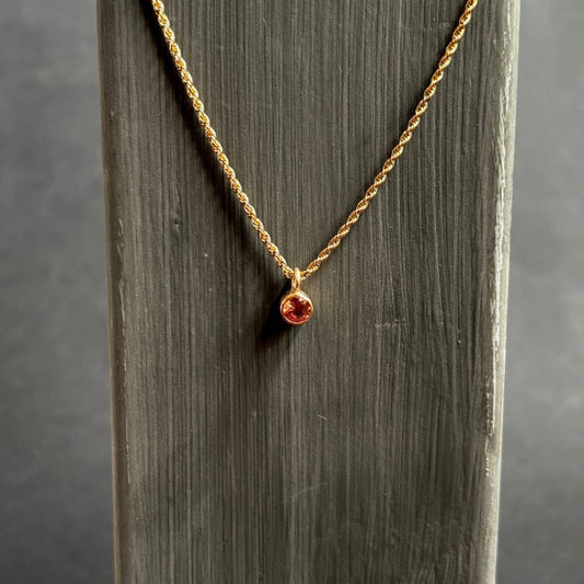 A delicate gold chain necklace with a small round amber-brown gemstone pendant, displayed on a vertical gray wooden stand against a dark gray background.