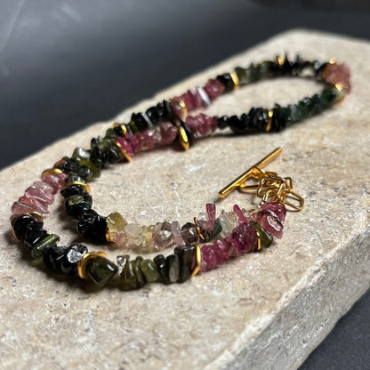 A beaded charm bracelet made of polished chunky gemstone beads in pink, green, and black, accented with gold spacer rings and a gold toggle clasp, displayed on a textured light gray stone slab.