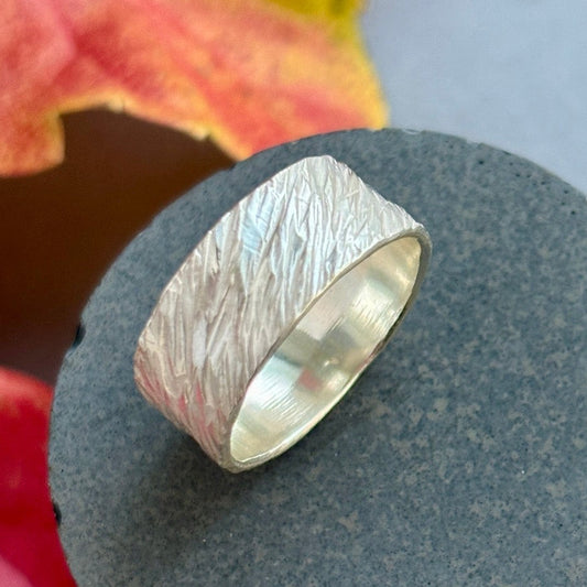 Silver ring with a textured surface on a dark stone surface with autumn leaves in the background