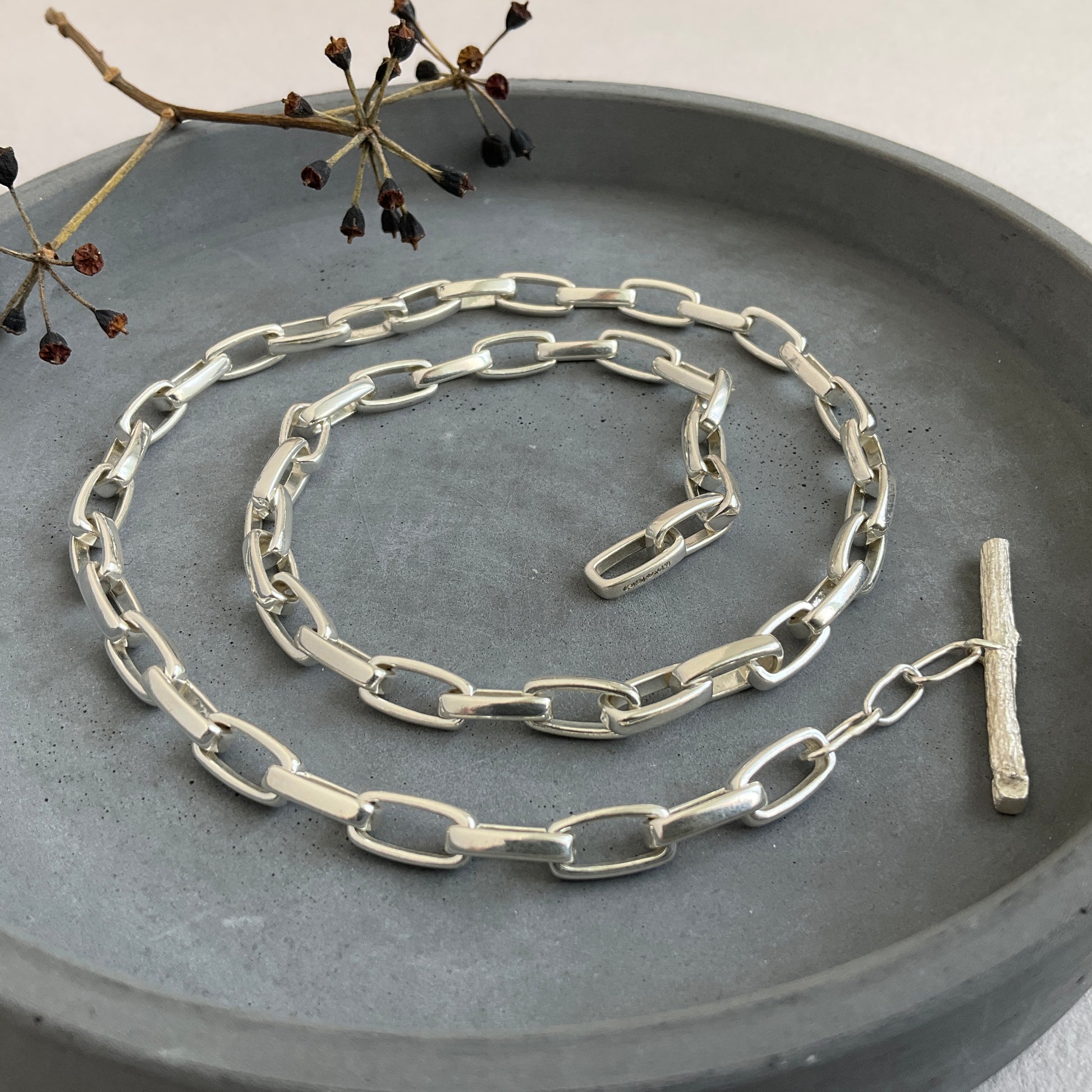 Tierra collection chunky chain in recycled sterling silver with mono casted twig clasp hallmarked by London Assay office. Displayed in a grey round dish with some black seeds on the left side 