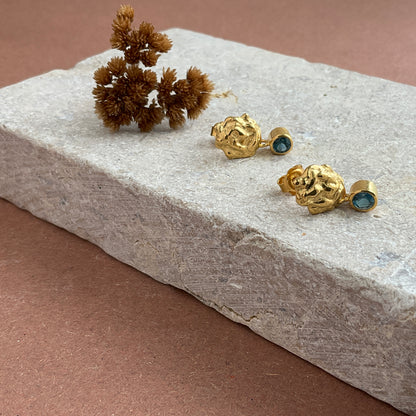 A pair of gold-colored drop earrings with blue topaz gemstones, displayed on a textured grey surface.