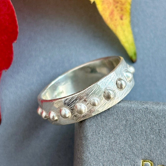 Silver ring with textured design on a gray surface with a red flower and yellow leaf.