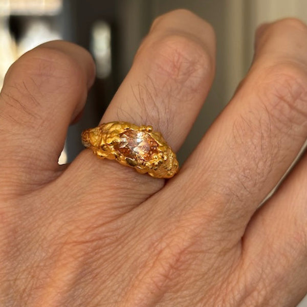 A close-up of a hand wearing a chunky, organic-shaped gold ring with a textured, hammered surface.