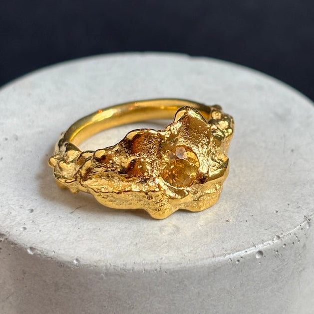 A gold ring with an organic, textured, rough stone-like design, displayed on a round white concrete pedestal against a dark background.