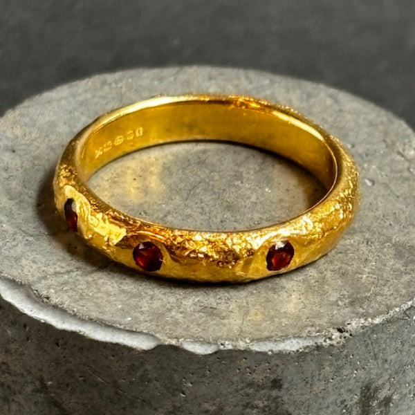 A textured gold ring with small red gemstone accents, displayed on a round gray stone pedestal against a dark gray background.