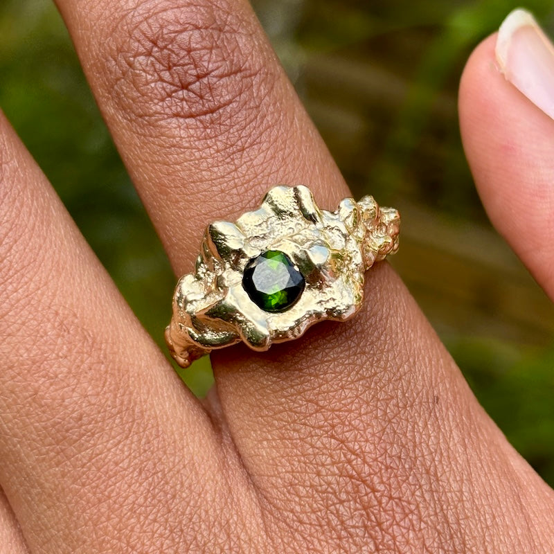 A close-up of a textured gold ring with a central green gemstone, worn on a finger against a blurred green background.
