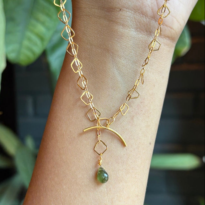 A gold chain necklace with a geometric link design, a crescent moon pendant, and a small green teardrop stone, shown worn on a person's wrist against a blurred background of green leaves.