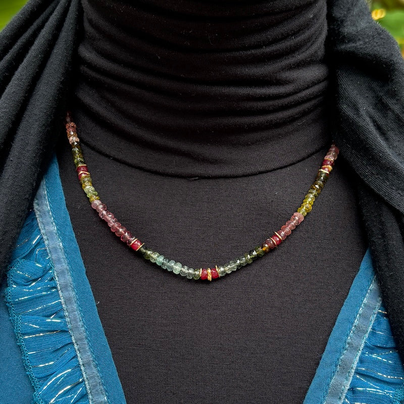A beaded necklace with small faceted gemstone beads in green, red, and yellow tones, displayed on a black turtleneck sweater worn by a model.