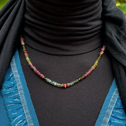 A beaded necklace with small faceted gemstone beads in green, red, and yellow tones, displayed on a black turtleneck sweater worn by a model.
