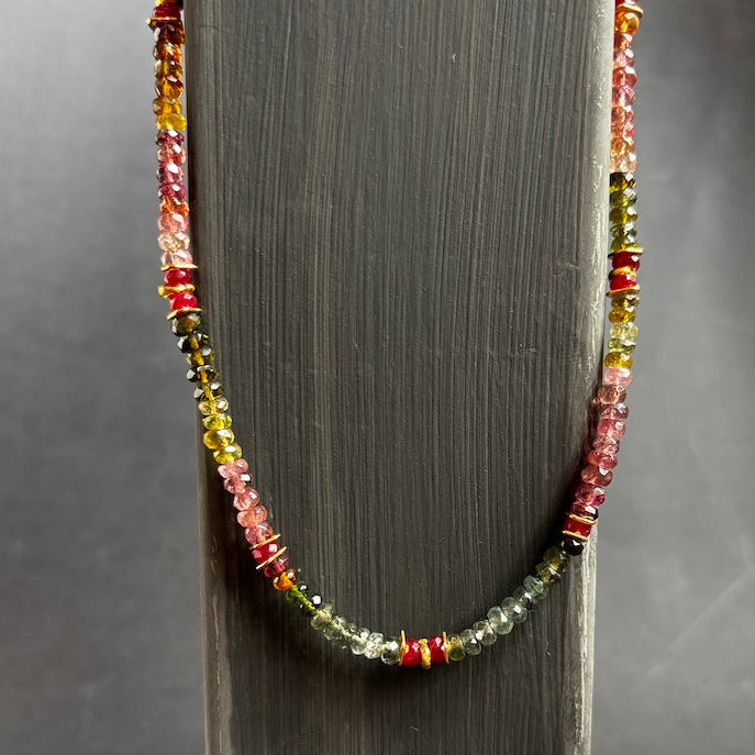 A beaded necklace draped over a dark gray wooden display stand, featuring faceted beads in pink, yellow, green, and red tones.