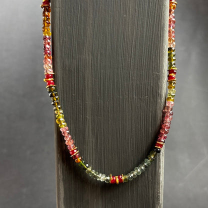 A beaded necklace draped over a dark gray wooden display stand, featuring faceted beads in pink, yellow, green, and red tones.