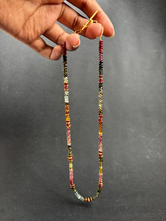 A beaded necklace with small multicolored gemstone-like beads and gold-toned clasp, held by a hand against a dark gray background.