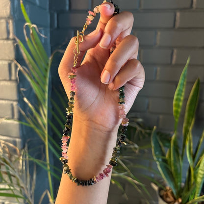 A hand holding a beaded necklace made of polished pink, green, and black gemstone chips with gold-toned metal accents, set against a dark gray brick wall and green plants.
