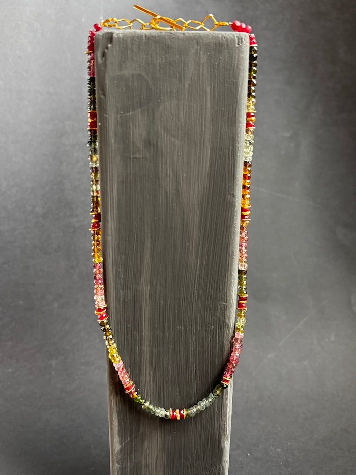 A beaded necklace with small multicolored beads in red, yellow, green, and orange, displayed on a dark gray wooden stand against a dark gray background.