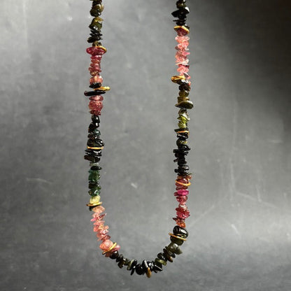A beaded necklace made of small, irregular polished gemstone chips in pink, black, green, and gold, suspended against a dark gray gradient background.