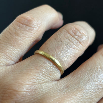 A close-up of a hand wearing a simple textured yellow gold band ring against a dark background.