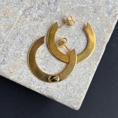 A pair of gold crescent-shaped hoop earrings with a small oval black and gold stone set in the front, displayed on a textured light gray stone slab against a dark background.
