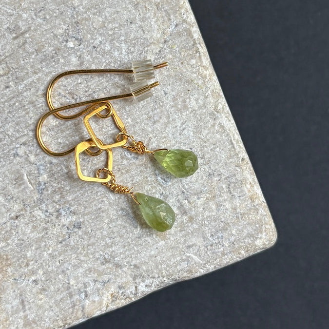 A pair of gold drop earrings with geometric wireframe tops and faceted green gemstone pendants, displayed on a textured gray stone slab against a dark background.