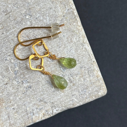 A pair of gold drop earrings with geometric wireframe tops and faceted green gemstone pendants, displayed on a textured gray stone slab against a dark background.