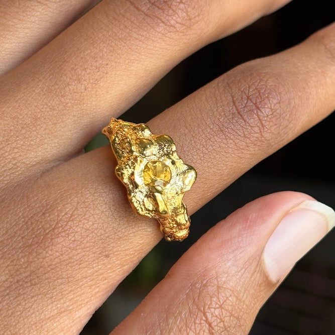 A close-up of a hand wearing a chunky, textured gold ring with a rough, organic shape.