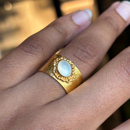 A close-up of a hand wearing a wide gold ring with a round white cabochon stone in a textured bezel setting.