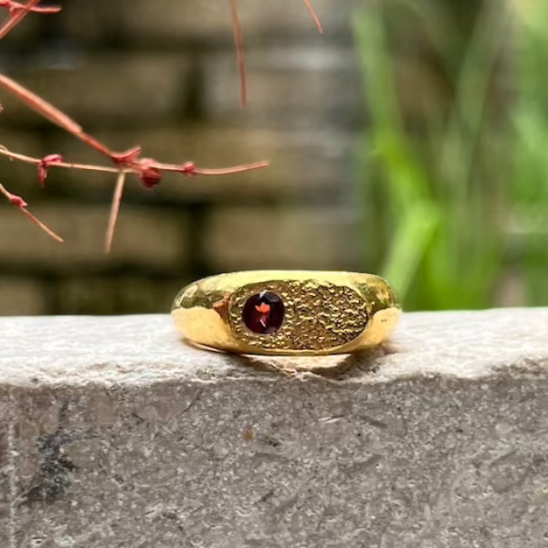 A textured gold ring with a central round red gemstone, resting on a gray stone ledge with a blurred outdoor background.