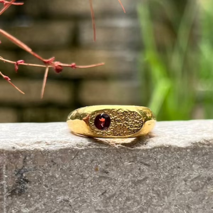 A textured gold ring with a central round red gemstone, resting on a gray stone ledge with a blurred outdoor background.