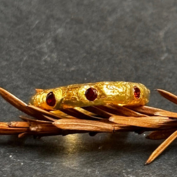 A textured gold ring with three small round red gemstone accents, displayed on a sprig of brown pine needles against a dark gray slate background.