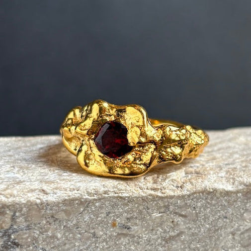 A gold ring with a textured, organic-shaped setting and a central dark red gemstone, resting on a light gray stone surface against a dark gray background.