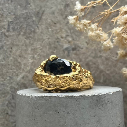 A textured gold ring with a faceted black gemstone, displayed on a round concrete pedestal with dried flowers in the background.